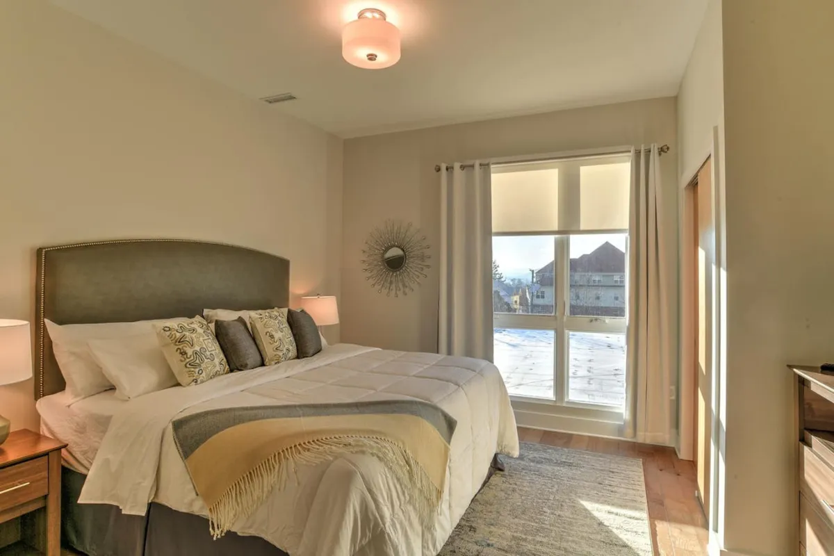 This image features a cozy bedroom with a king-size bed, flanked by nightstands, a window letting in soft natural light, a mirror, and a plush rug. The calming color palette and neatly made bed with quality linens evoke a sense of peace and relaxation, making it a perfect depiction of a serene retreat.