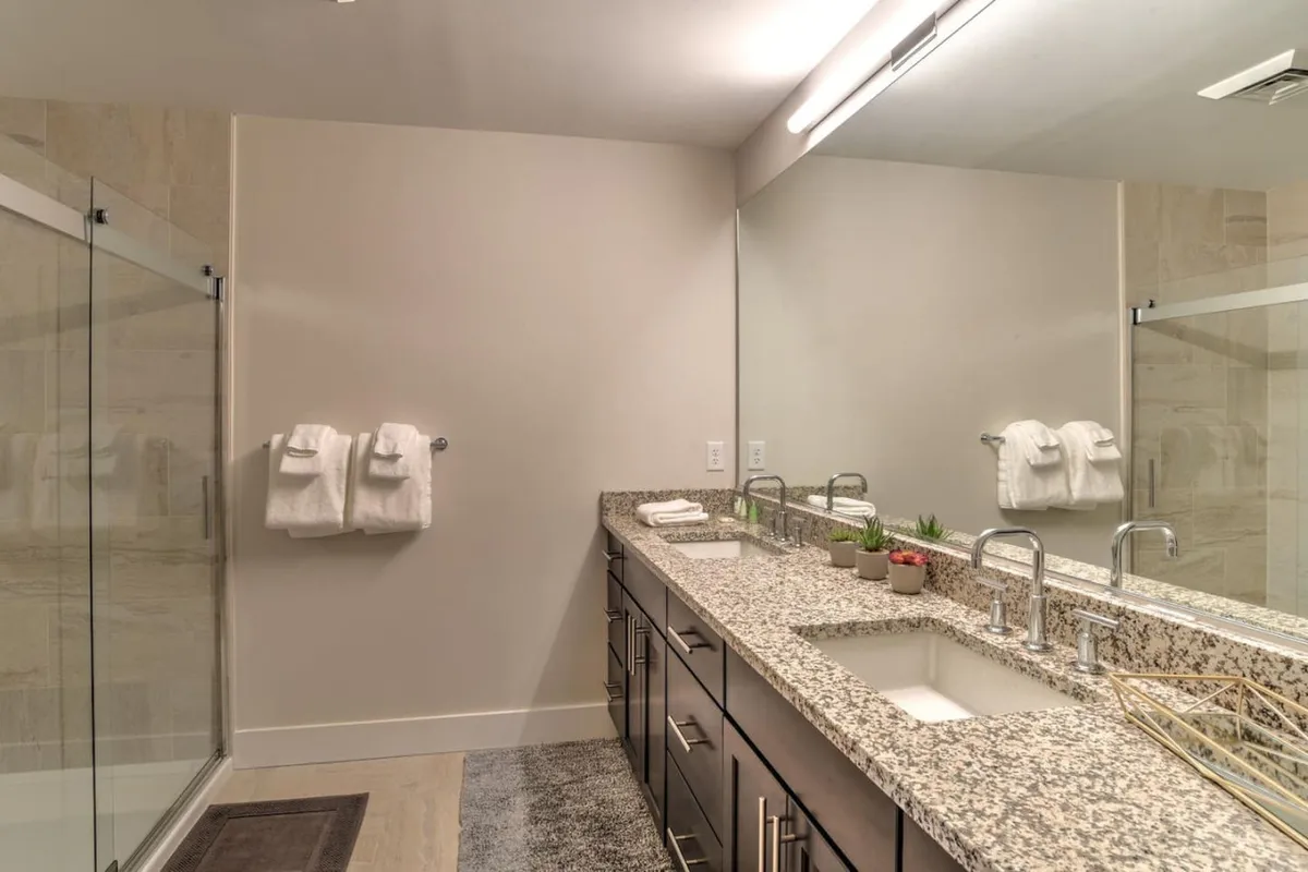 This modern bathroom scene showcases a granite countertop with double vanity sinks and a large mirror. The space is well-lit and includes a glass shower enclosure, fluffy towels, and stylish fixtures, embodying both convenience and luxury.