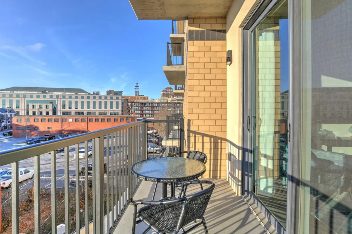 A spacious balcony with a glass table and chairs overlooks the cityscape and surrounding buildings, perfect for enjoying coffee or watching the sunset over the city. The relaxed urban vibe highlights the balcony as an intimate vantage point for special moments.