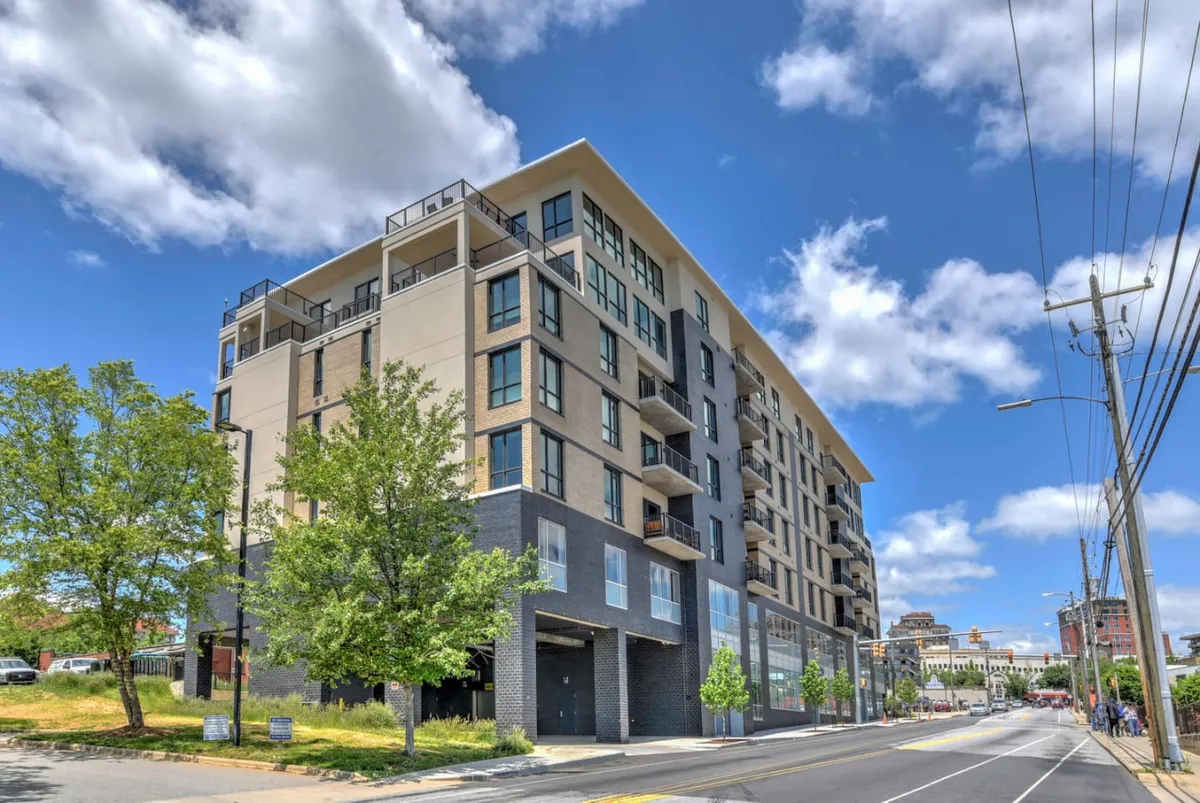 A dynamic exterior shot of the condo building showcases modern architecture with clean lines, stylish balconies, and lush green landscaping. The scene captures a vibrant urban atmosphere, hinting at proximity to city attractions and community spaces.