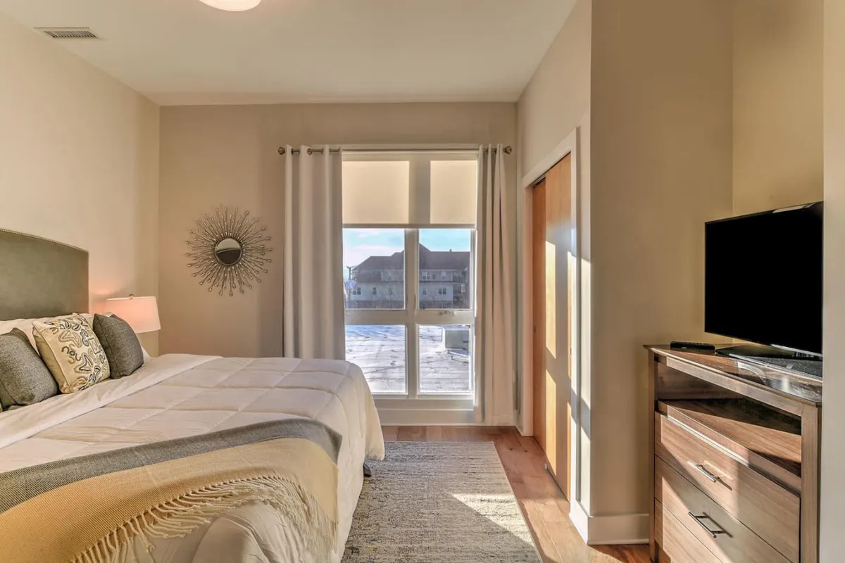 A serene bedroom with a large window letting in gentle sunlight, a comfortable bed adorned with decorative pillows, and a sleek, modern television unit. The room feels airy, uncluttered, and inviting, ideal for unwinding after a busy day.