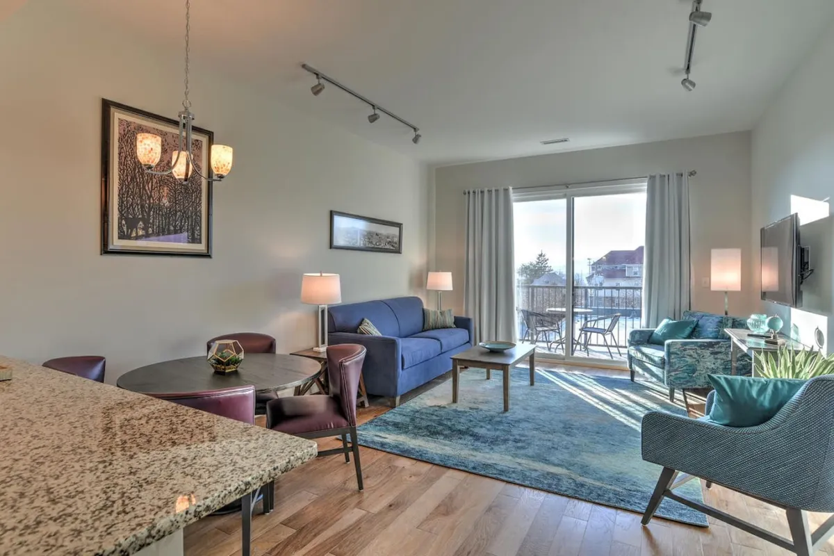 This image presents a welcoming living and dining space, with a plush sofa and a dining table positioned to take advantage of abundant natural light streaming through a large window. The room is styled with contemporary decor and offers a soothing atmosphere ideal for relaxation or entertaining.