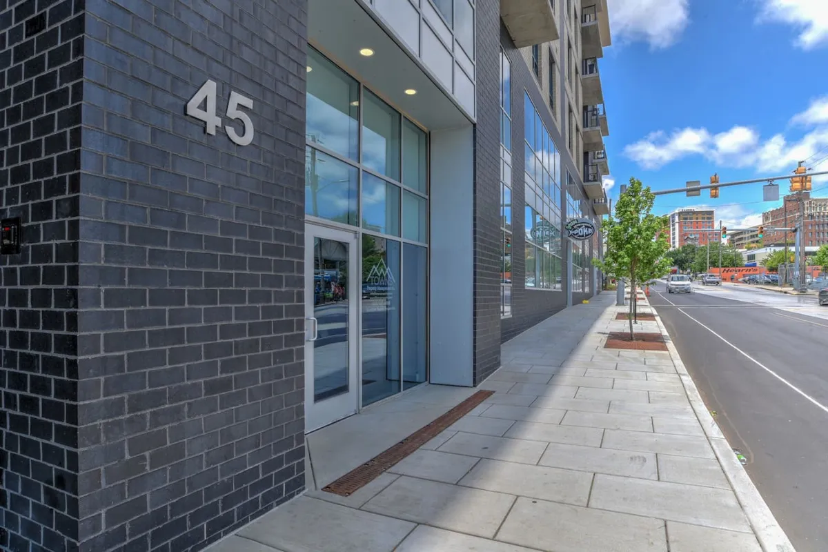 The image captures the building's exterior, focusing on the facade, the entrance door, street signage, and a nearby tree. There is a sense of movement and activity, suggesting a secure and welcoming entry point for residents and guests.