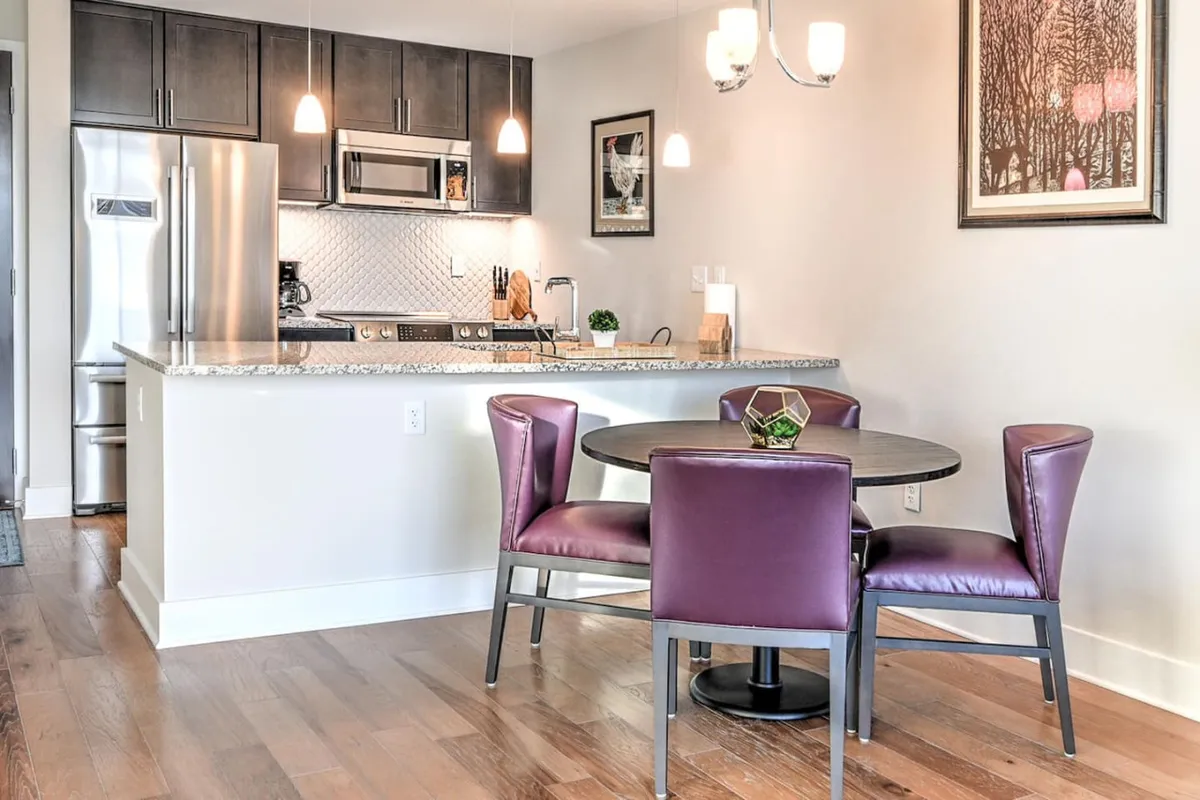 The kitchen island with striking purple chairs commands attention, complemented by a stylish dining table and vibrant wall art. The open-plan design merges cooking and social spaces, inviting both cozy family dinners and lively group meals. The lighting and décor lend a touch of sophistication and warmth.